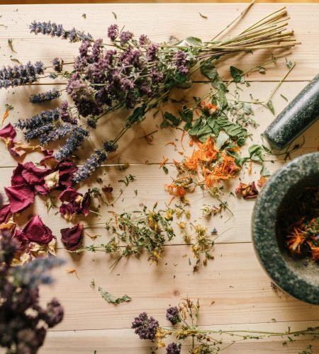 Flat lay of dried herbs and flowers with a stone mortar and pestle on a rustic wooden table.